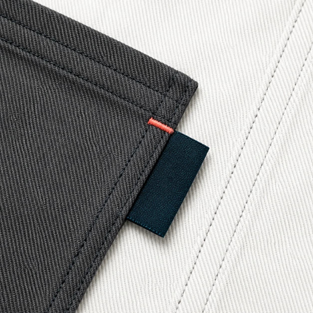A close study of a graphite garment pocket, ivory twill seam, and a narrow teal woven label with coral stitching.